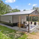 Modern post frame metal garage building with pewter gray sidewalls, two white roll-up doors, and a black roof, set on a concrete pad and surrounded by trees