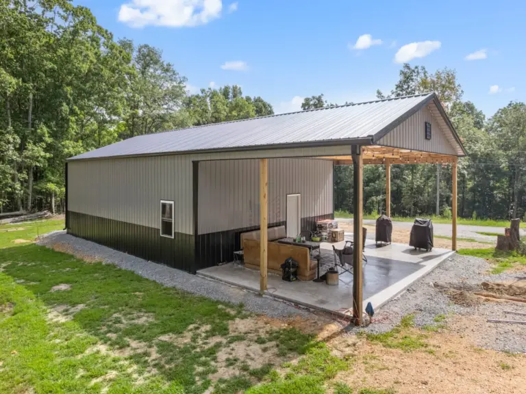 Modern post frame metal garage building with pewter gray sidewalls, two white roll-up doors, and a black roof, set on a concrete pad and surrounded by trees