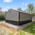 Modern post frame metal garage building with pewter gray sidewalls, two white roll-up doors, and a black roof, set on a concrete pad and surrounded by trees
