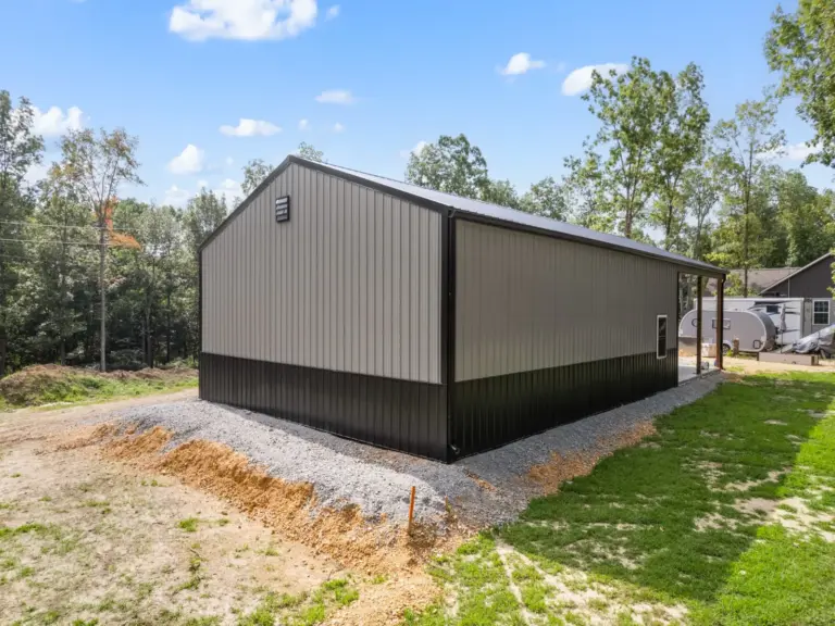 Modern post frame metal garage building with pewter gray sidewalls, two white roll-up doors, and a black roof, set on a concrete pad and surrounded by trees