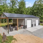Modern post frame metal garage building with pewter gray sidewalls, two white roll-up doors, and a black roof, set on a concrete pad and surrounded by trees