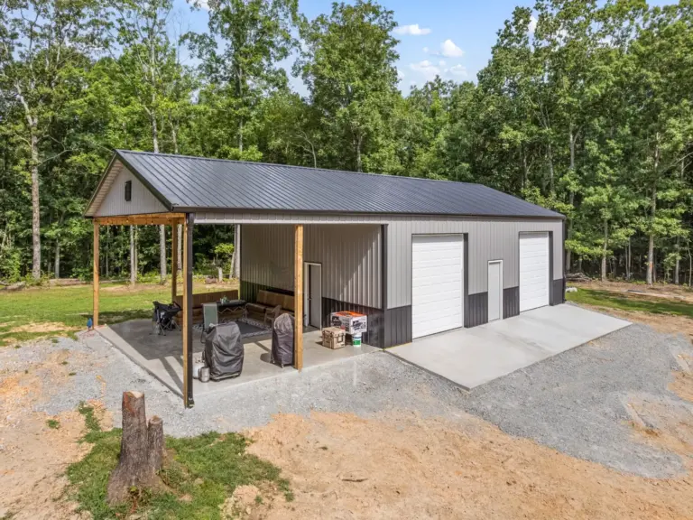 Modern post frame metal garage building with pewter gray sidewalls, two white roll-up doors, and a black roof, set on a concrete pad and surrounded by trees
