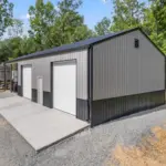 Modern post frame metal garage building with pewter gray sidewalls, two white roll-up doors, and a black roof, set on a concrete pad and surrounded by trees