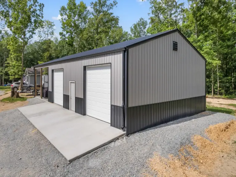 Modern post frame metal garage building with pewter gray sidewalls, two white roll-up doors, and a black roof, set on a concrete pad and surrounded by trees