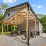 Modern post frame metal garage building with pewter gray sidewalls, two white roll-up doors, and a black roof, set on a concrete pad and surrounded by trees
