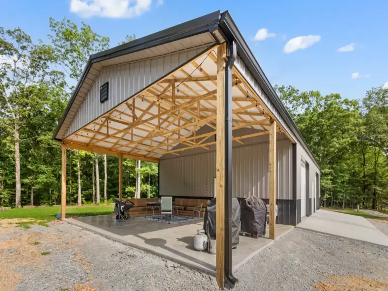 Modern post frame metal garage building with pewter gray sidewalls, two white roll-up doors, and a black roof, set on a concrete pad and surrounded by trees