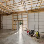 Interior of a post frame metal garage with exposed trusses, two large roll-up garage doors, concrete floor, and workshop equipment including fans and scaffolding