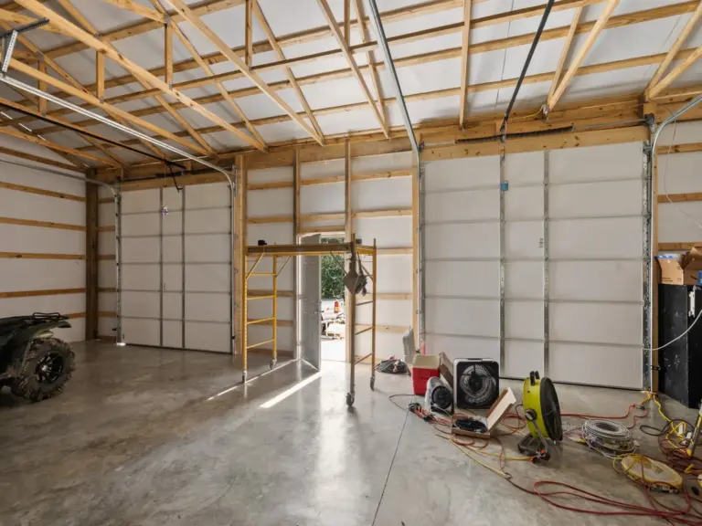 Interior of a post frame metal garage with exposed trusses, two large roll-up garage doors, concrete floor, and workshop equipment including fans and scaffolding