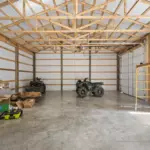 Spacious interior of a post frame garage with exposed wood trusses, concrete floor, overhead garage doors, and ATVs and tools arranged in a functional workshop setting