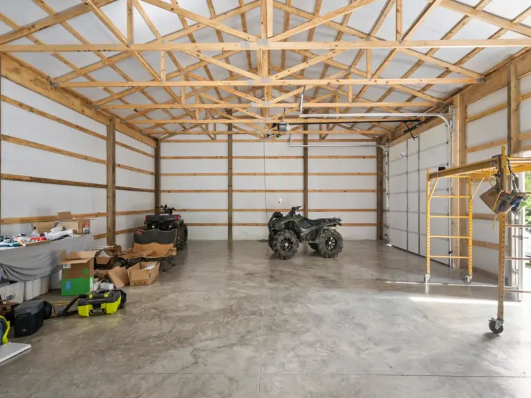 Spacious interior of a post frame garage with exposed wood trusses, concrete floor, overhead garage doors, and ATVs and tools arranged in a functional workshop setting