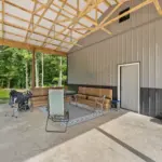 Covered outdoor living space attached to a post frame metal building, featuring a concrete floor, exposed trusses, patio seating, and wooded surroundings