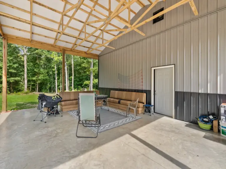 Covered outdoor living space attached to a post frame metal building, featuring a concrete floor, exposed trusses, patio seating, and wooded surroundings